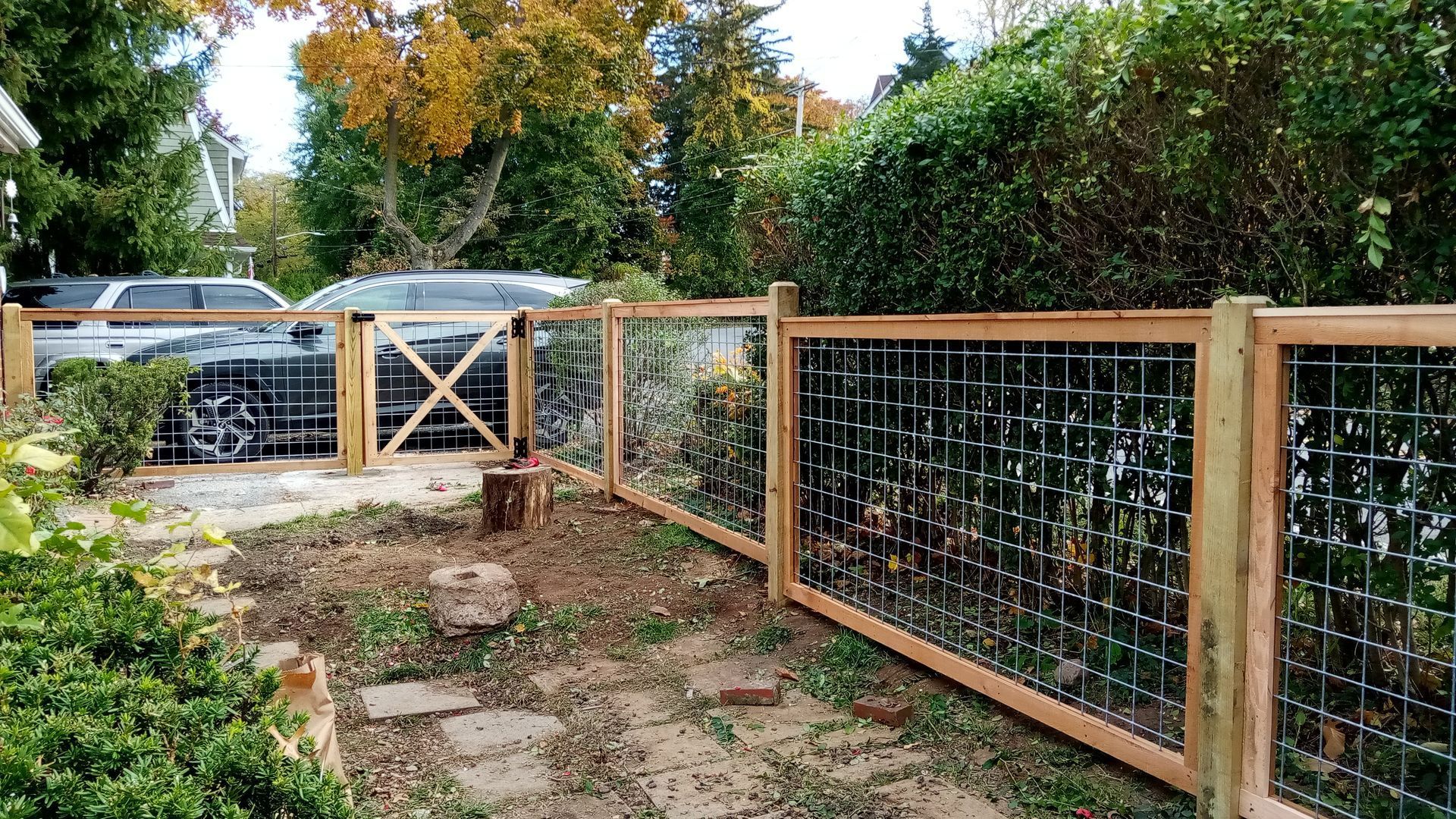 Wooden fence with wire mesh panels enclosing a yard, gate visible. Cars and trees in the background.