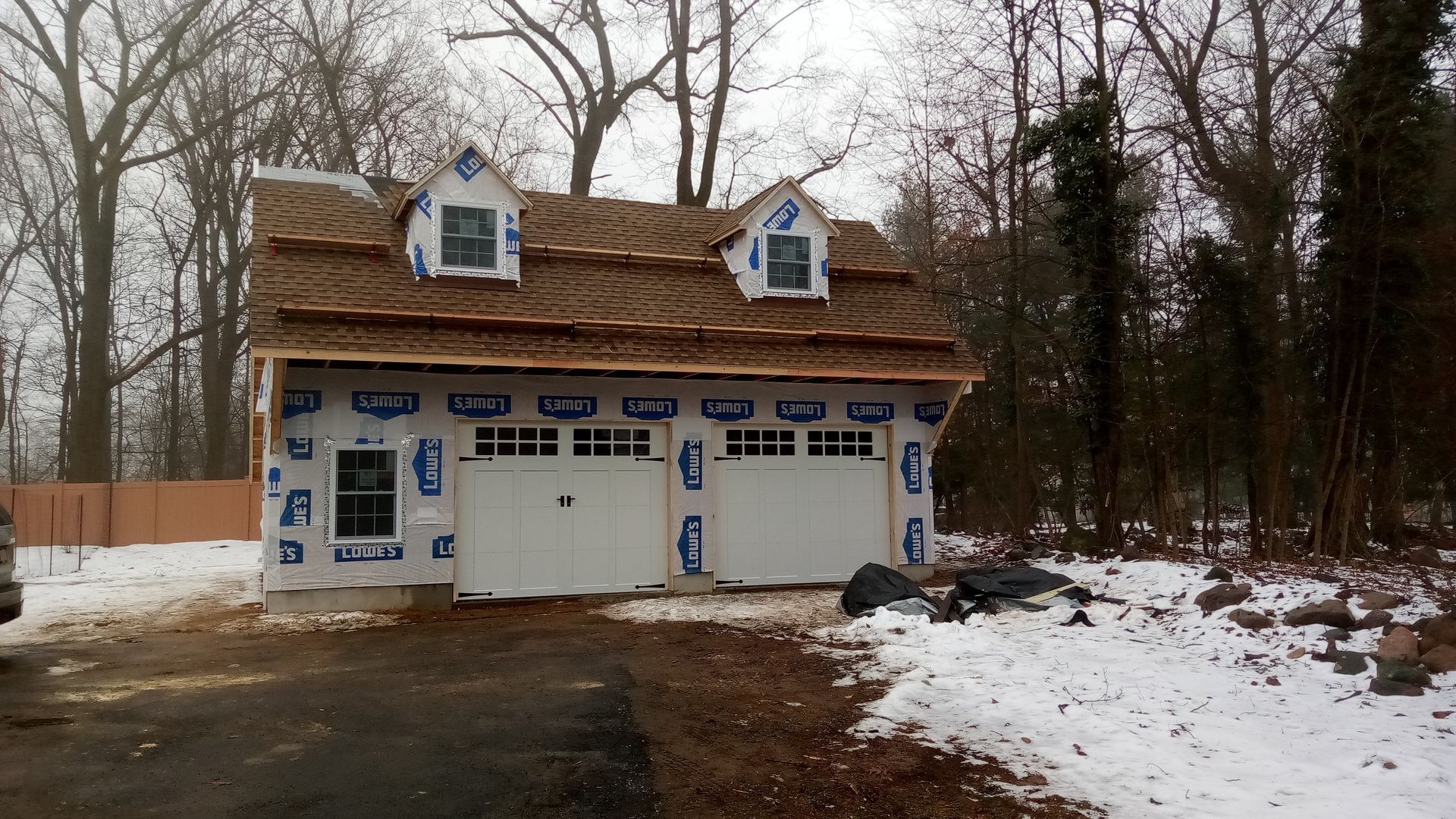 Garage under construction, two white doors, two dormers, brown roof, snowy ground, trees in the background.
