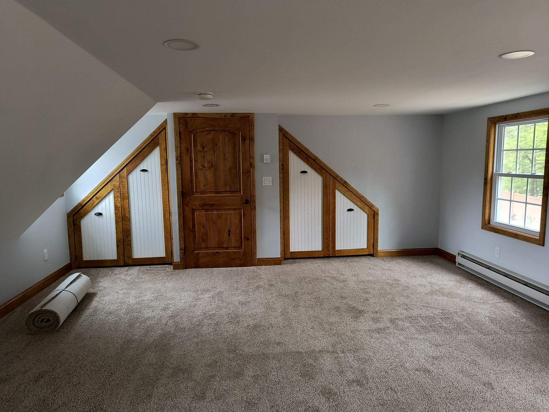 Interior of a room with carpet, slanted walls, built-in storage, and a window with a radiator.