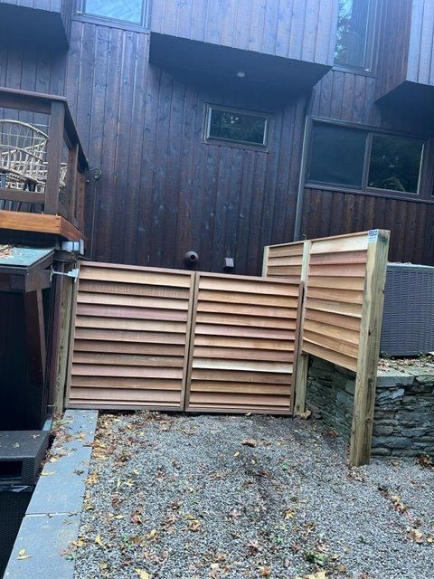 Wooden slatted gate with cedar siding house exterior.  Asphalt and gravel ground; some leaves.