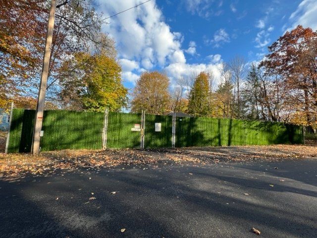 Green artificial hedge fence bordering a road under a blue sky with autumn trees.