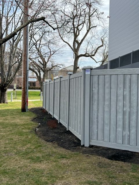 Gray vertical-slat fence along a grassy lawn, beside a building. Bare trees and a utility pole are visible in the background.