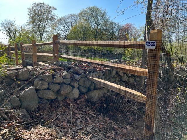 Wooden split-rail fence with wire mesh attached, atop a stone wall, in a sunny outdoor setting.