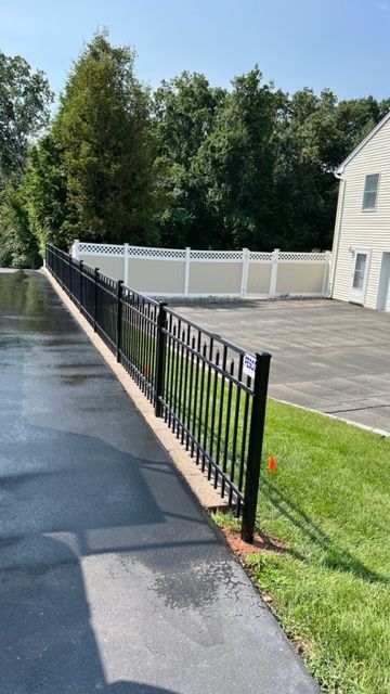 Black metal fence bordering a driveway, with white fence in background and house, trees.