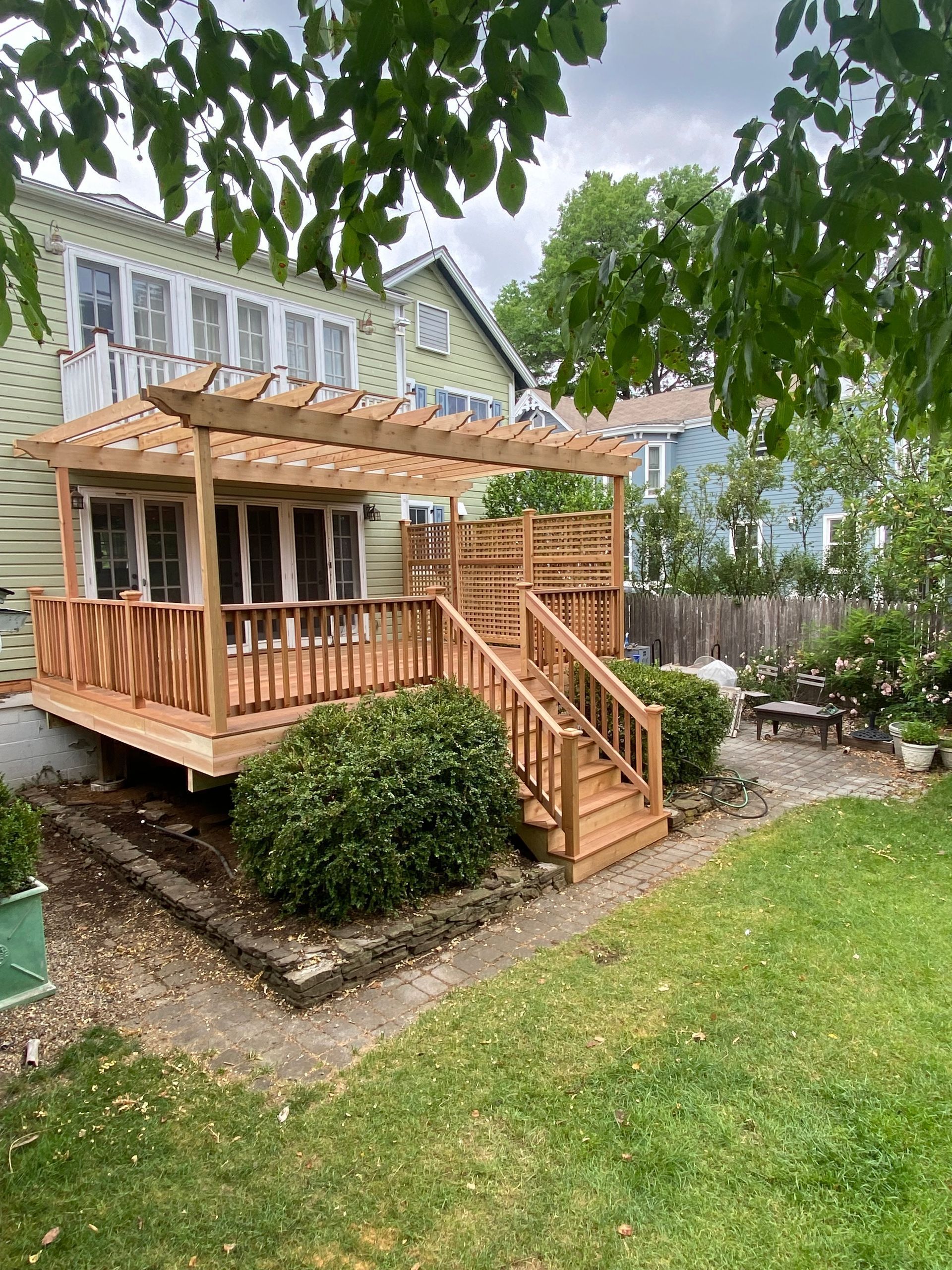 Wooden deck with pergola, stairs, and privacy screen in a backyard with greenery and a bench.