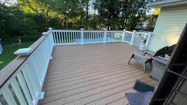 Brown wooden deck with white railing, overlooking a yard and trees. Two chairs are on the deck.