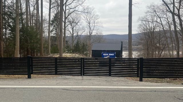 Black horizontal slatted fence with gate across a road, behind a dark building, trees, and hills.