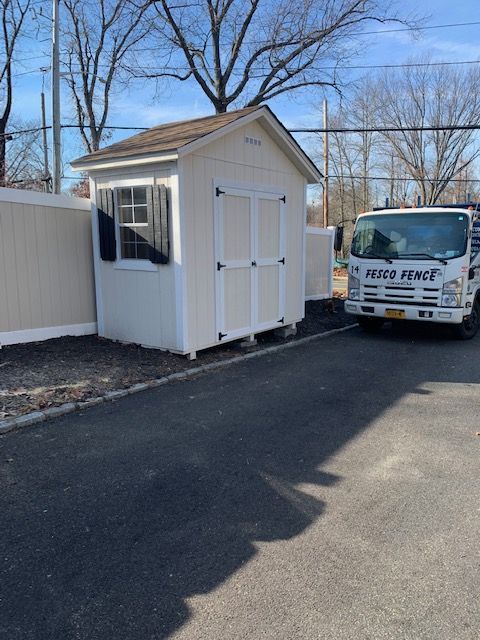 A shed being delivered by a truck on a paved driveway next to a fence.