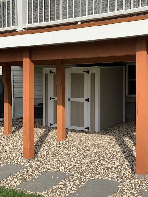 Two tan storage sheds under a deck with brown support beams and gravel ground.