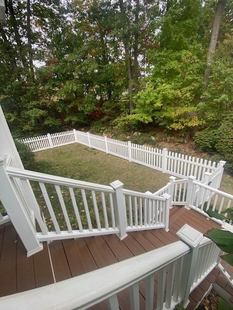 White picket fence encloses grassy yard viewed from a brown wooden deck with white railing. Trees in background.