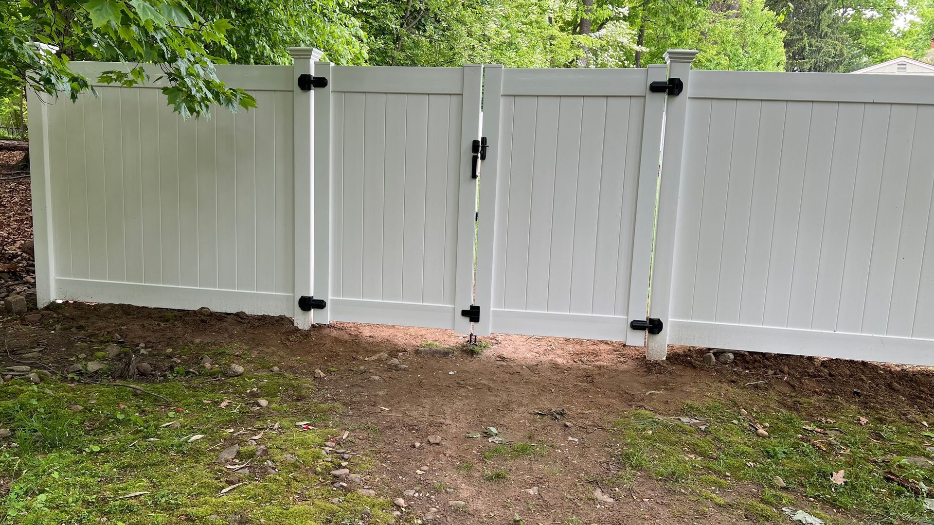 White vinyl fence with gate, black hardware. Green grass and brown soil in the foreground. Trees in background.
