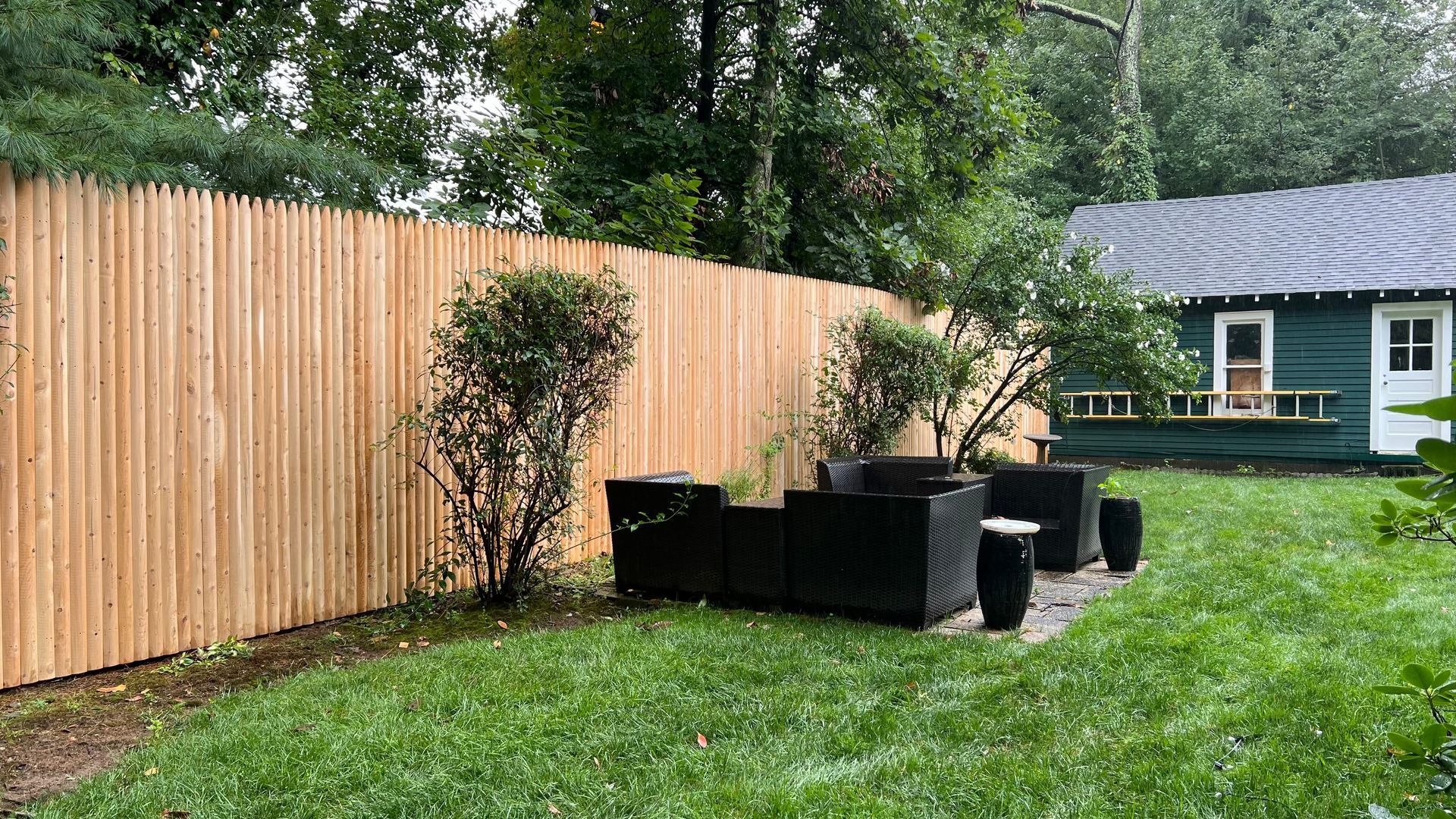 Wooden fence in backyard with patio furniture and small green bushes. Green grass and a blue house.
