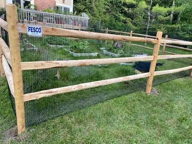 Wooden split-rail fence with black netting around a garden on green grass.