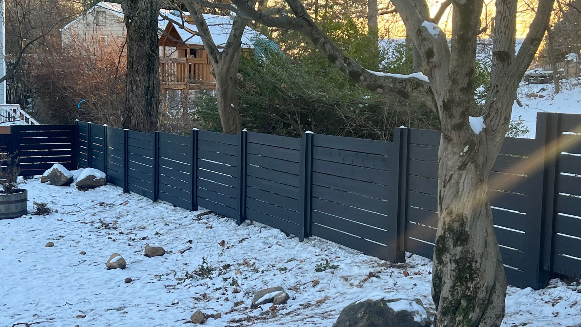 Dark gray horizontal slat fence in snow-covered yard with a tree and distant house.