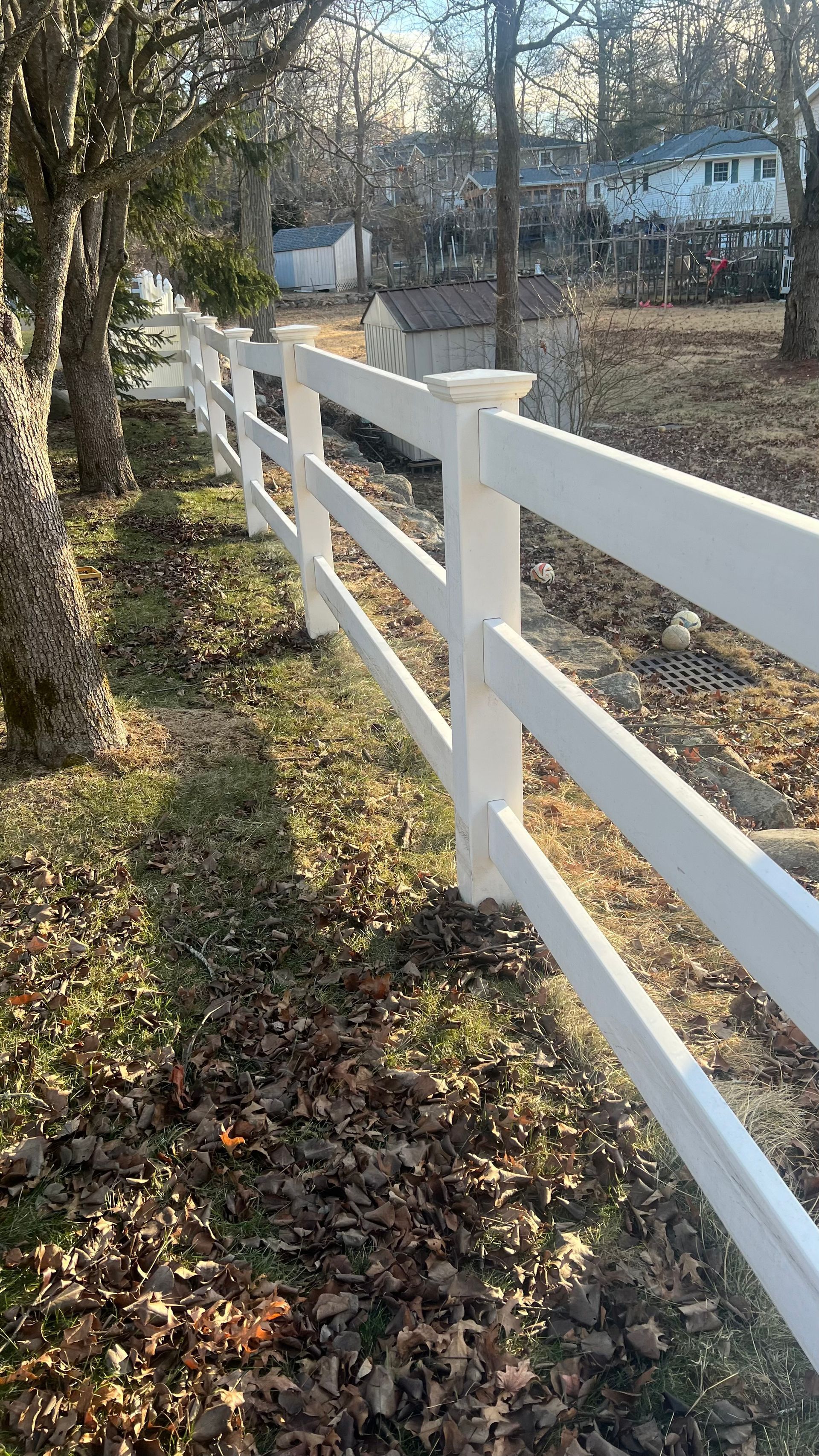 White three-rail fence in a grassy yard, with trees and houses visible in the background on a sunny day.