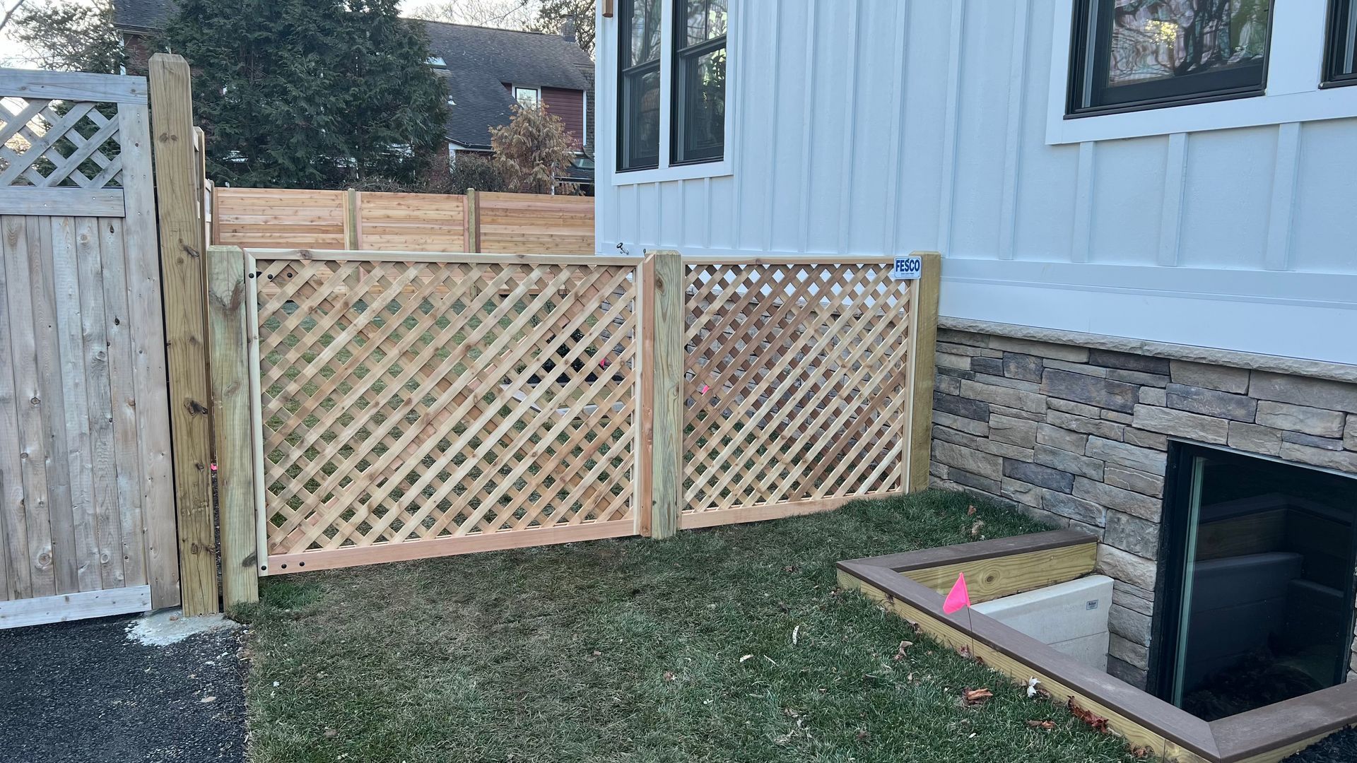 Wooden lattice fence and gate next to a house with a basement access, in a yard.