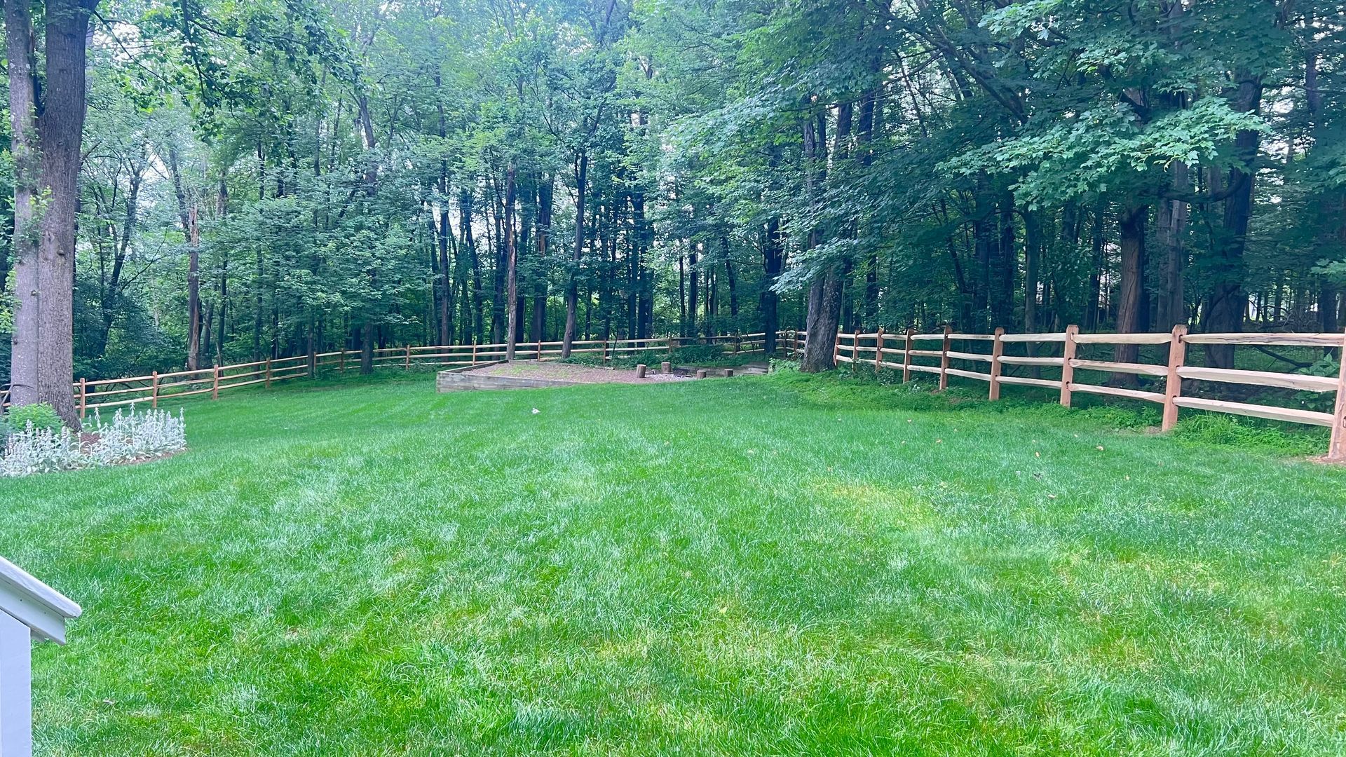 Green lawn enclosed by split-rail fence, leading to a wooded area with mature trees under an overcast sky.