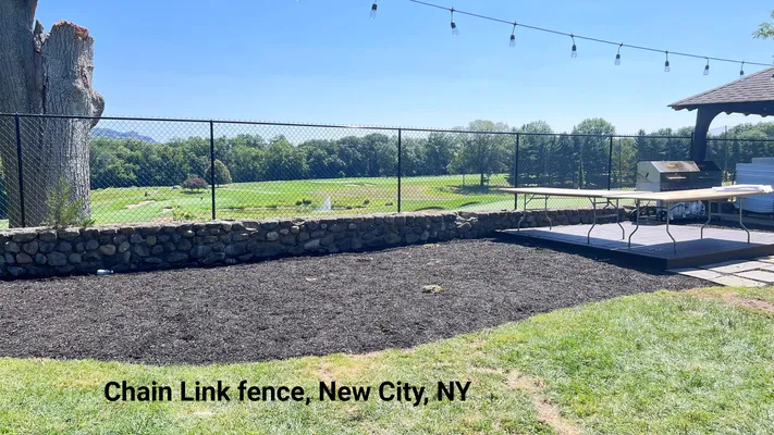 Black chain link fence bordering a grassy area with a view of green fields, trees, and sky in New City, NY.