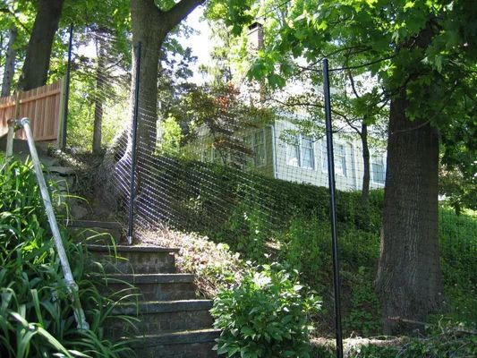 Stairs ascend a grassy hill next to a chain-link fence, leading toward a house. Trees surround the scene.