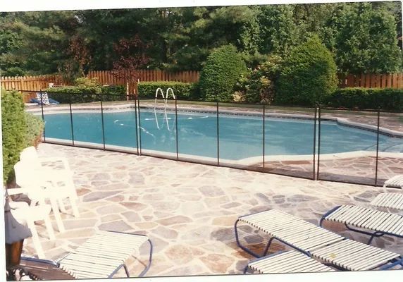 Pool surrounded by a black mesh safety fence. Stone patio with white chairs and lounge chairs. Trees in the background.