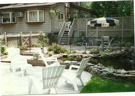 Backyard with pond, patio, white chairs, lattice fence, and multi-level deck, under a house.