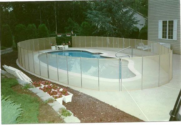 Pool with a beige safety fence surrounding it. Concrete patio, house, and bushes in the background.