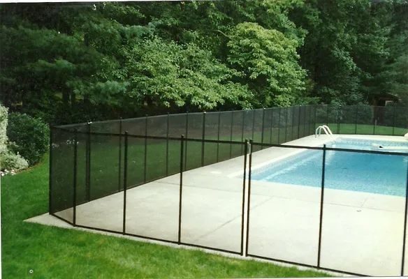 Black mesh pool safety fence surrounds a swimming pool on a concrete deck, with greenery in the background.