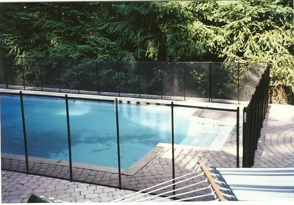 Black mesh safety fence around a blue swimming pool in a brick patio, green trees in the background.