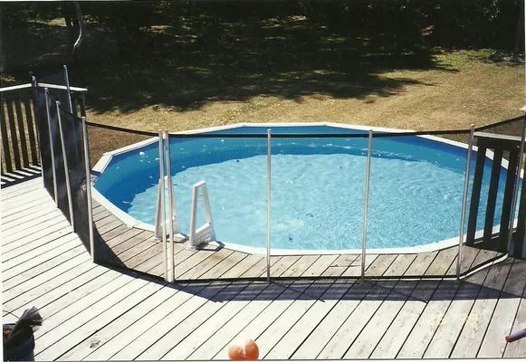 Above-ground pool with safety fence on wooden deck, surrounded by grass and trees.