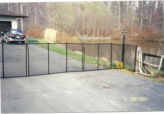 Black mesh safety fence by a pond, spanning a driveway. Car parked near a house.