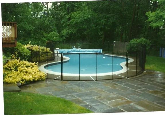 Pool with safety fence, surrounded by a stone patio and green foliage. A covered pool is surrounded by a black fence.