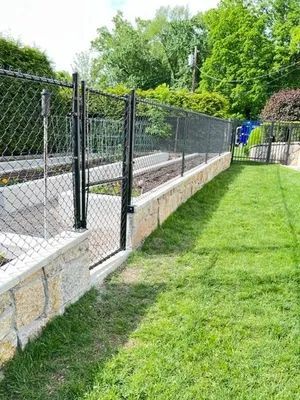 Chain-link fence with gate atop a stone wall, bordering a grassy area and garden beds.