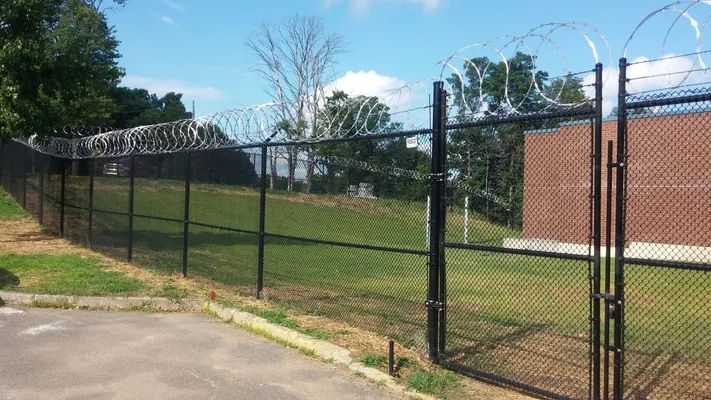 Black chain-link fence topped with razor wire, enclosing a grassy area and brick building under a blue sky.
