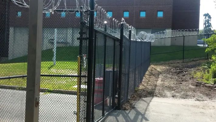 Black security fence with razor wire, enclosing a brick building with blue-tinted windows. Concrete walkway.