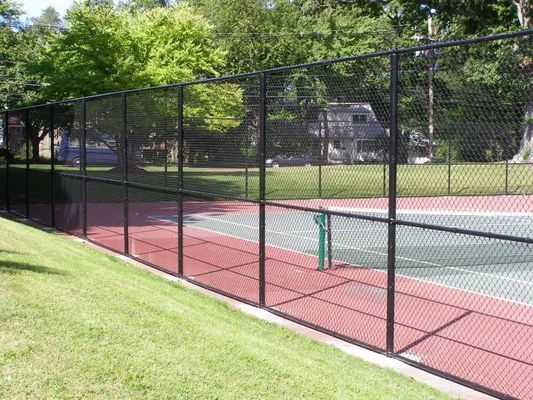 Black chain-link fence surrounding a tennis court with red and green surface. Trees in the background.