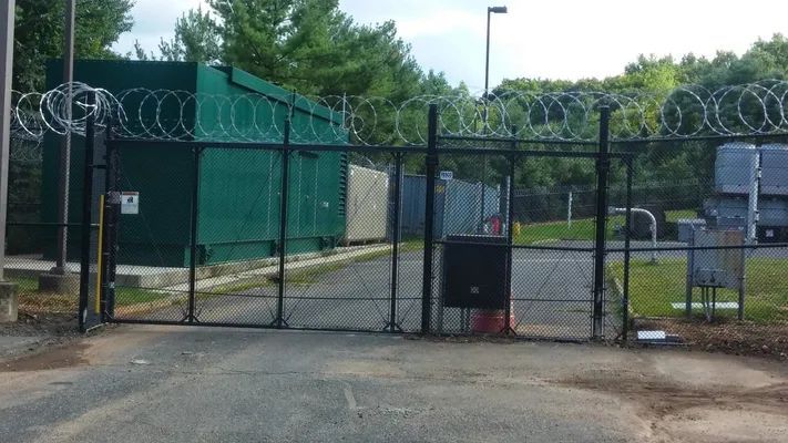 Black security gate topped with razor wire, enclosing green utility buildings.