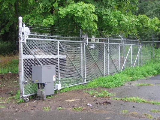 Sliding chain-link gate with barbed wire, gray motor box, and green grassy area in front.