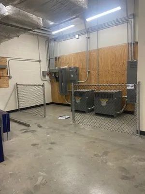 A fenced-in utility area with electrical boxes, conduits, and two gray units on a concrete floor.