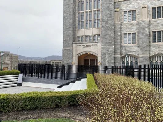 Stone building entrance with black fence, bushes, and a cloudy sky.