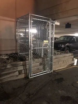 Chain-link fence enclosure in a parking garage. Contains mechanical equipment near a retaining wall. A vehicle is parked in the background.