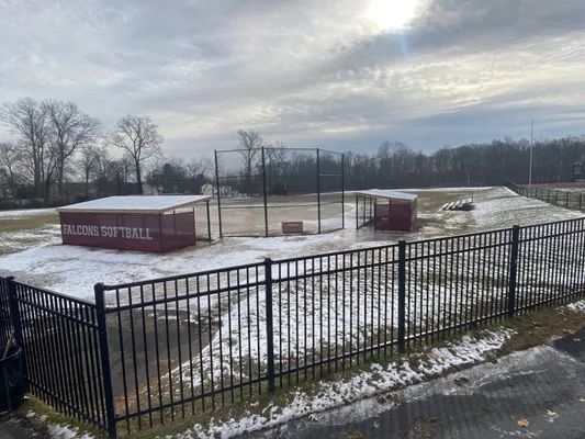 A softball field with snowy ground, a fence, dugouts, and a batting cage under a cloudy sky.