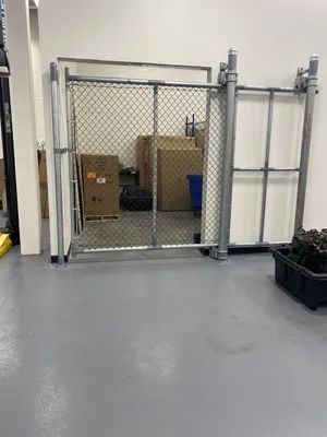 Chain-link gate inside a warehouse, guarding stored boxes and containers against a white wall. Gray floor.