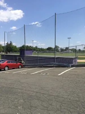 Baseball field with purple dugout, protective netting, and parked cars. Blue sky.