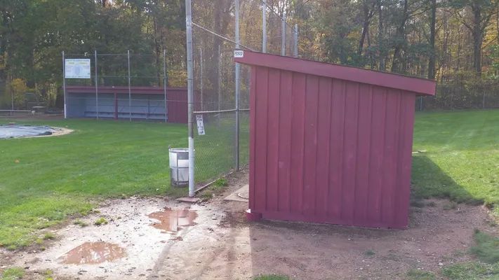 Red dugout beside a chain link fence and a baseball field with muddy ground.