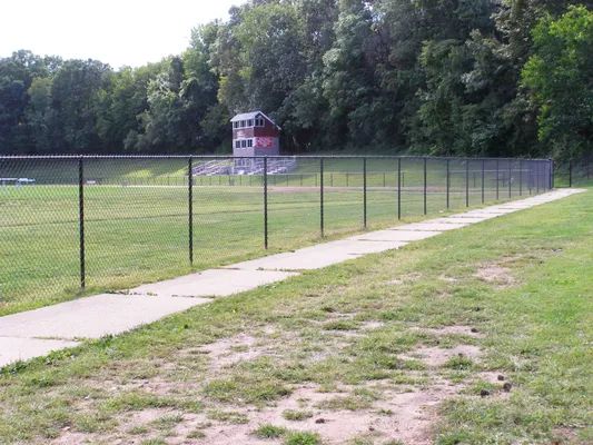 A chain-link fence and concrete path border a grassy field, with a small red building in the background.
