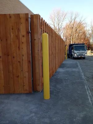 Yellow bollard next to a tall wooden fence and a truck on pavement.