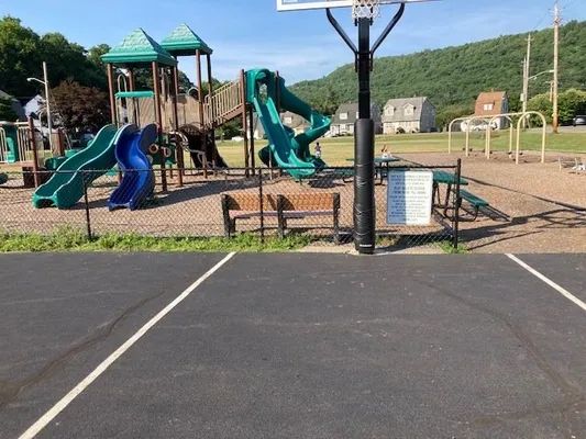 Playground with slides, swings, and basketball hoop, set against a green hill and sunny sky.