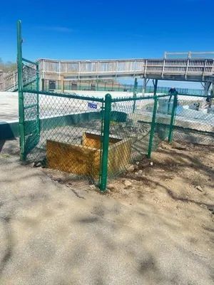 Green chain-link fence surrounds a wooden box on a sandy area, with a wooden walkway in the background.