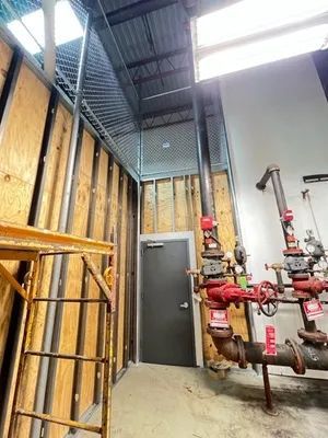 Interior view of a construction site. A ladder leads to a caged area above a door. Pipes and valves are visible.