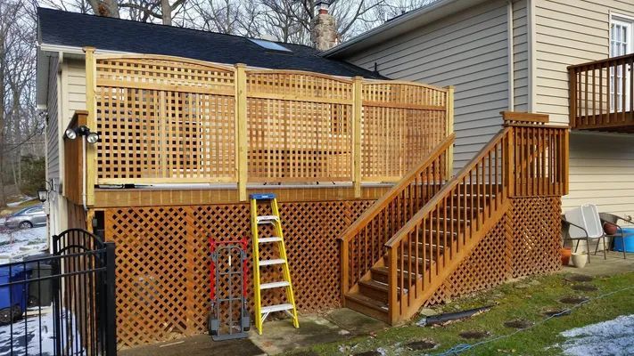 Wooden deck with lattice panels, stairs, and a ladder against a beige house with a dark roof.